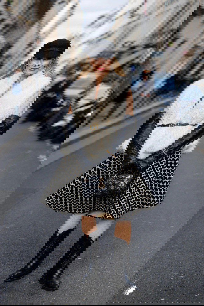 Стильная женщина в кремовой пушистой жилетке, зафиксированной ремнём, в юбке с геометрическим принтом и высоких сапогах — фотокадр уличной моды.