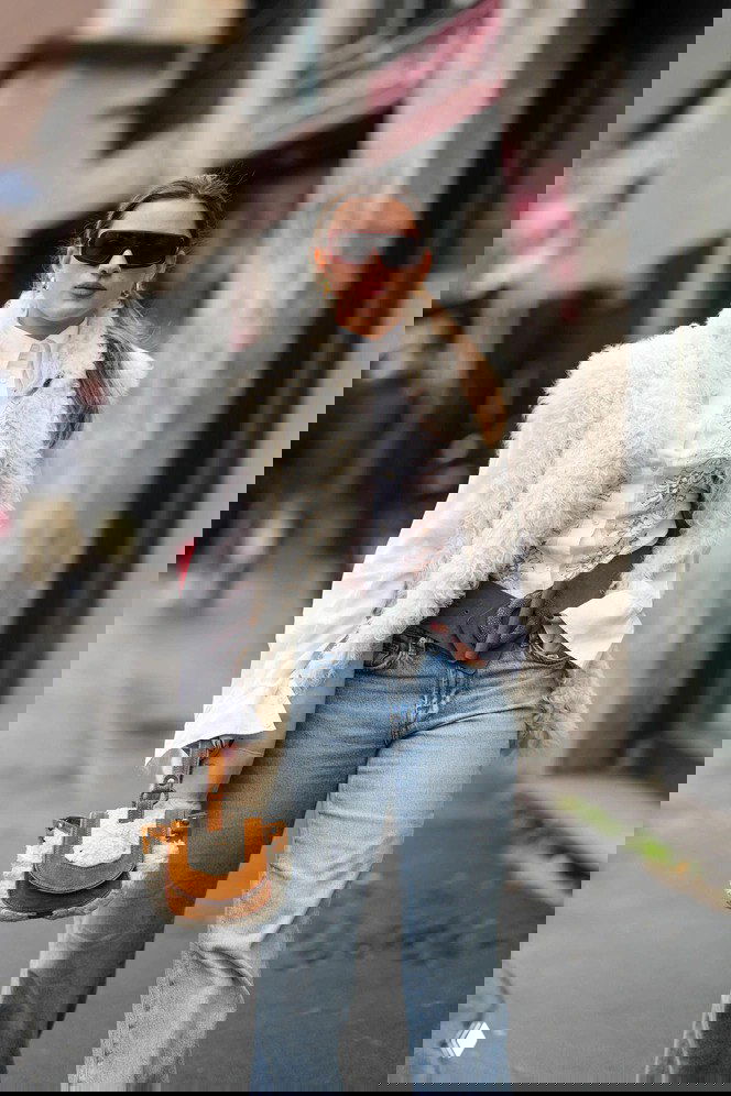 Молодая девушка в белой объёмной меховой жилетке и джинсах, с маленькой сумкой с меховой отделкой — спокойный уличный образ в стиле бохо.