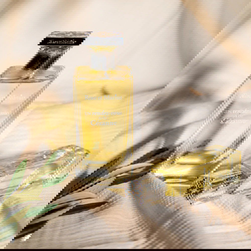 Флакон Rose et Marius — Un Rendez-vous Avec Cézanne на мягком текстильном фоне с оливковой веткой; кадр подчёркивает богатую кожано-хвойную структуру и пряные ноты композиции.