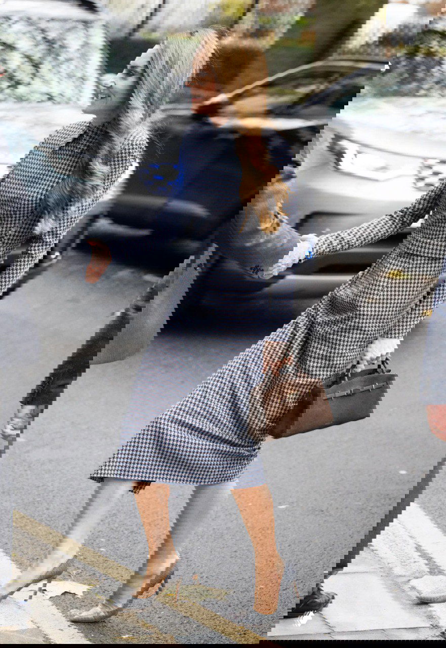 Фигура женщины в ломаной клетке идёт по тротуару в серых туфлях и держит коричневую сумку, уличная сцена с автомобилями на заднем плане