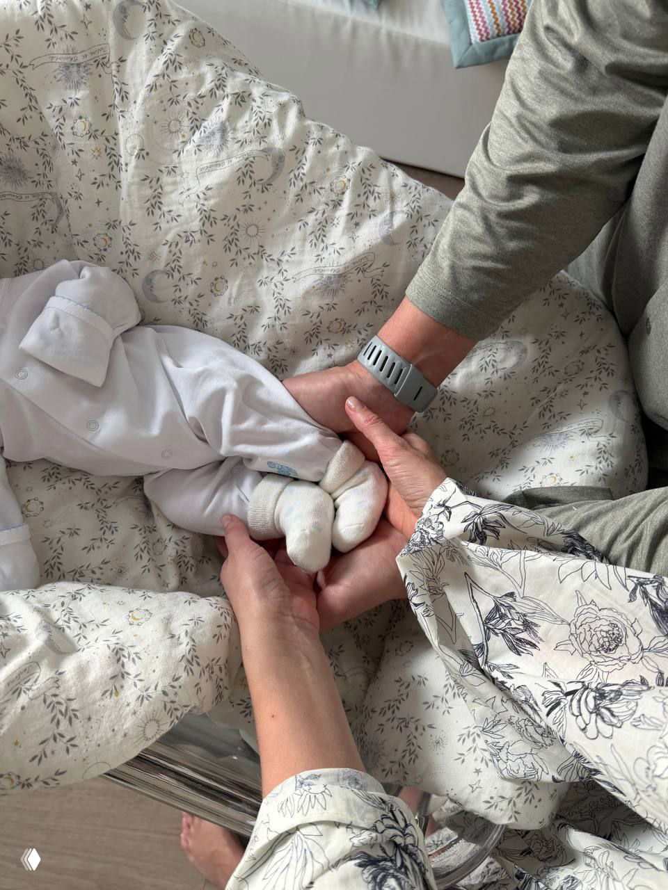 Фото в дома: несколько взрослых рук держат ножки новорождённого в белых носочках на фоне мягкой люльки и постельного белья с неброским узором.