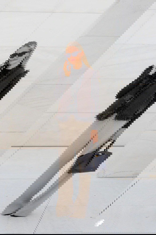 Блондинка в тёмной дублённой жилетке с меховой отделкой, светлые прямые брюки и чёрная сумка на фоне минималистичной архитектуры — элегантный образ.