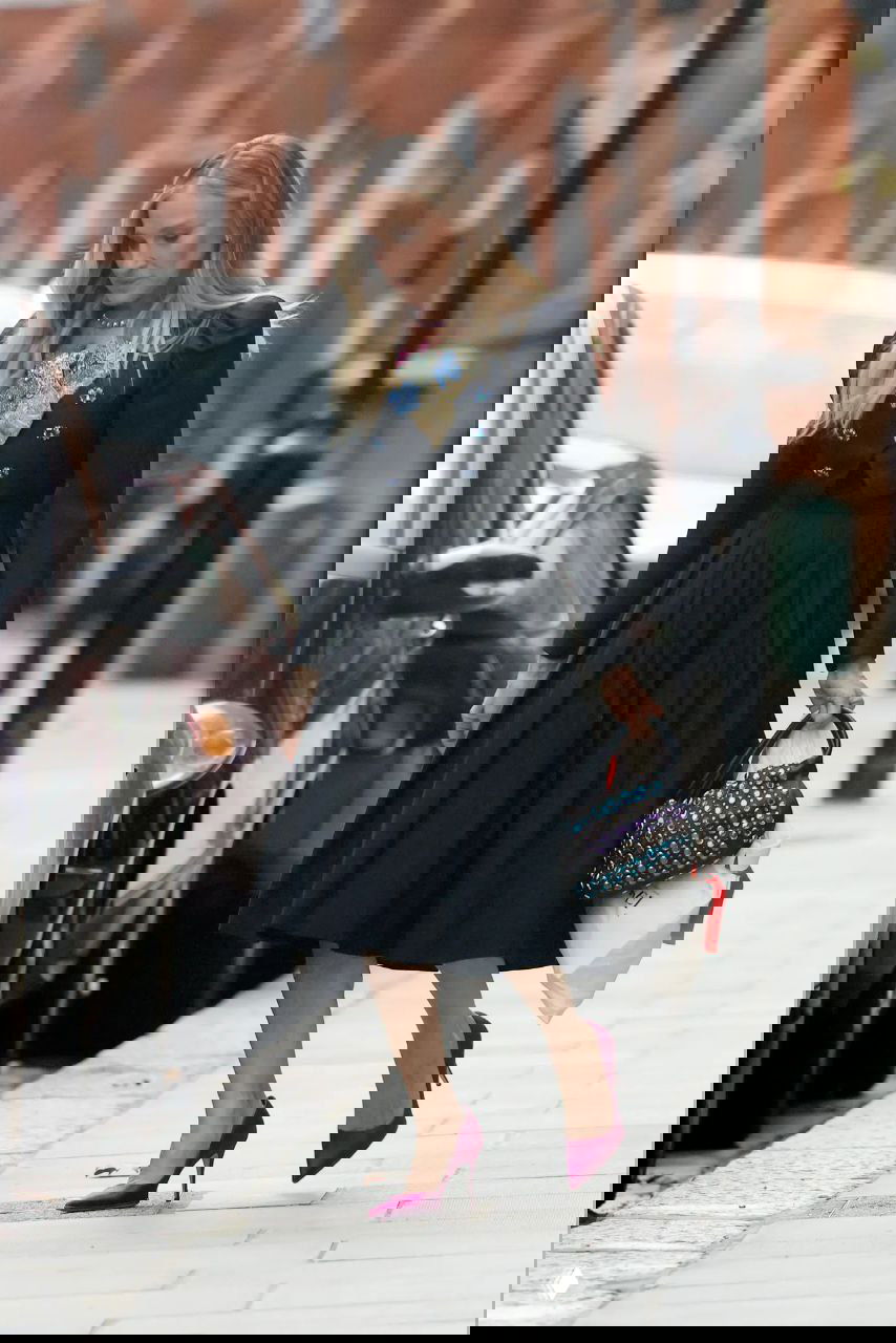 Сара Джессика Паркер в чёрном платье и бордовых туфлях идёт по лондонской улице рядом с местом проведения мероприятия, уличная сцена и автомобили.