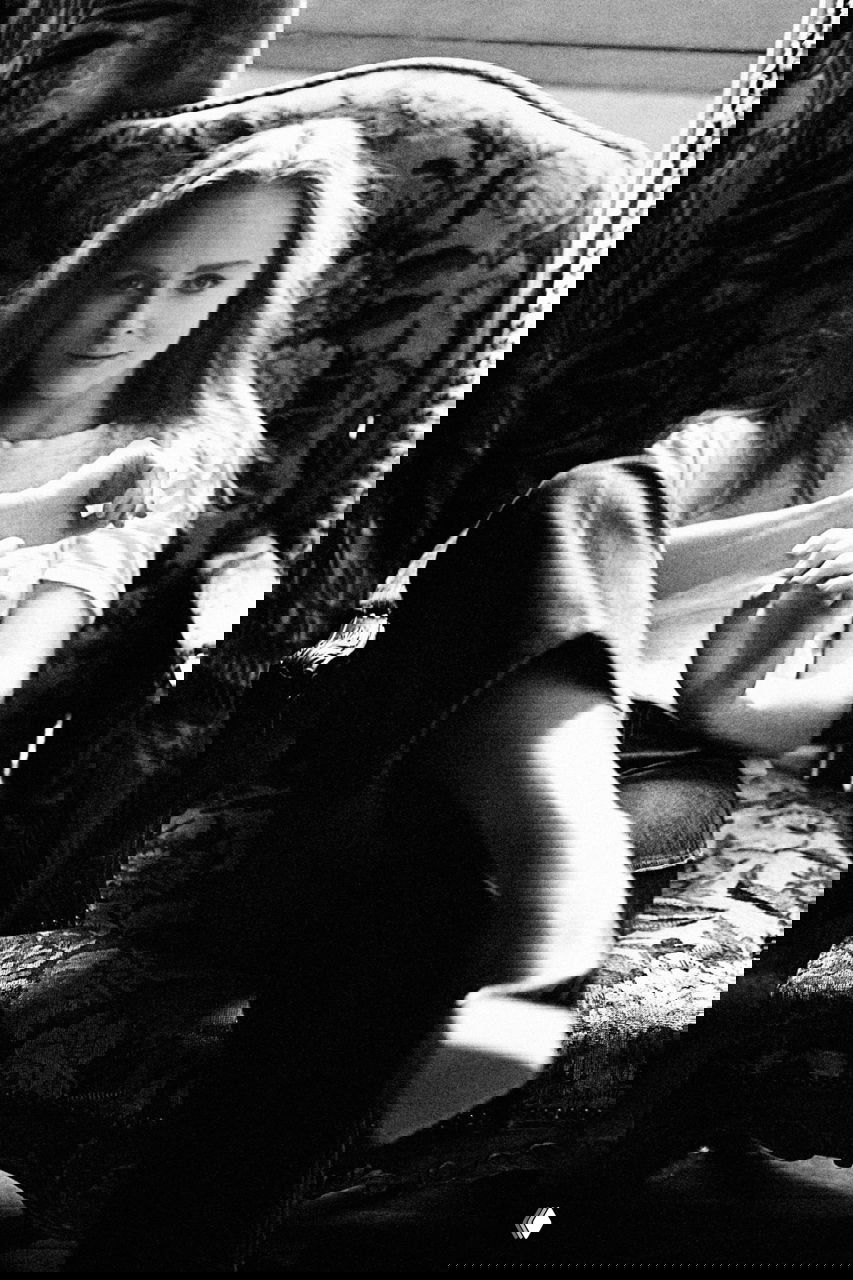 Чёрно‑белый портрет женщины в кресле; расслабленная поза, взгляд в камеру, винтажный интерьер и мягкое рассеянное освещение.