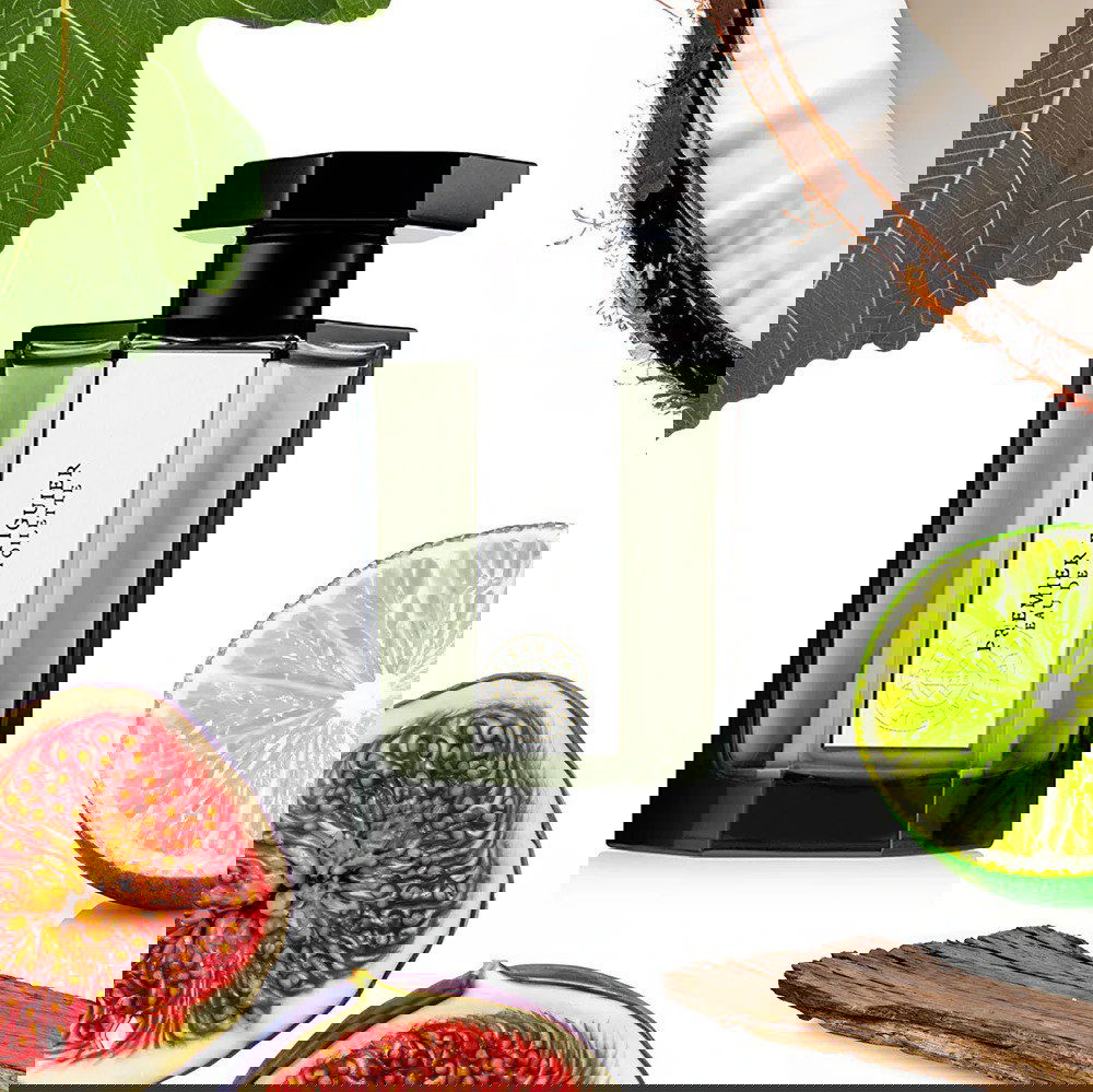 Флакон L’Artisan Parfumeur — Premier Figuier на светлом фоне с кусочками инжира и лайма; кадр передаёт зелёно-фруктовый старт и древесно-кокосовую базу аромата.