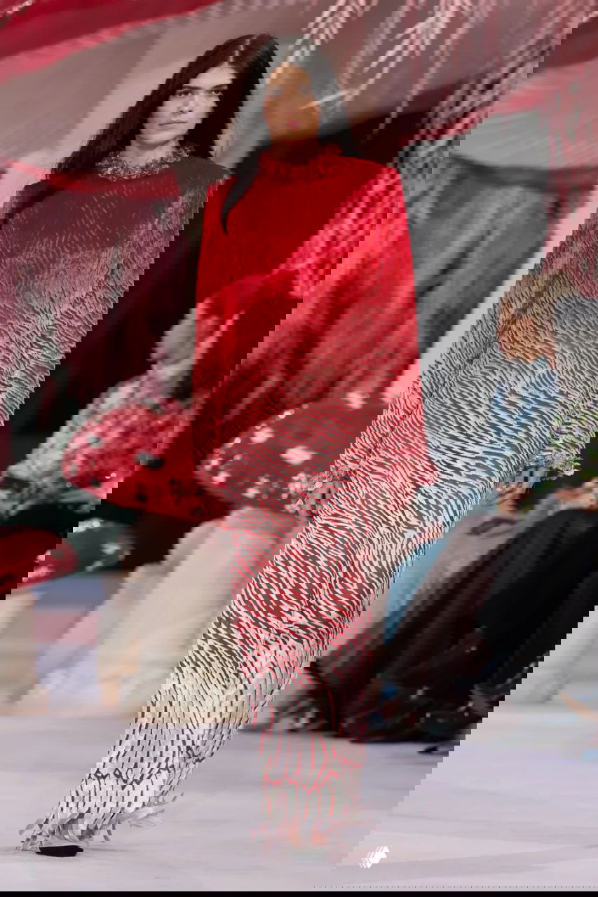 Модель в длинном красно-белом платье с зебровидным рисунком идёт по подиуму Chanel на фоне крупных декоративных грибов и сказочной сцены.