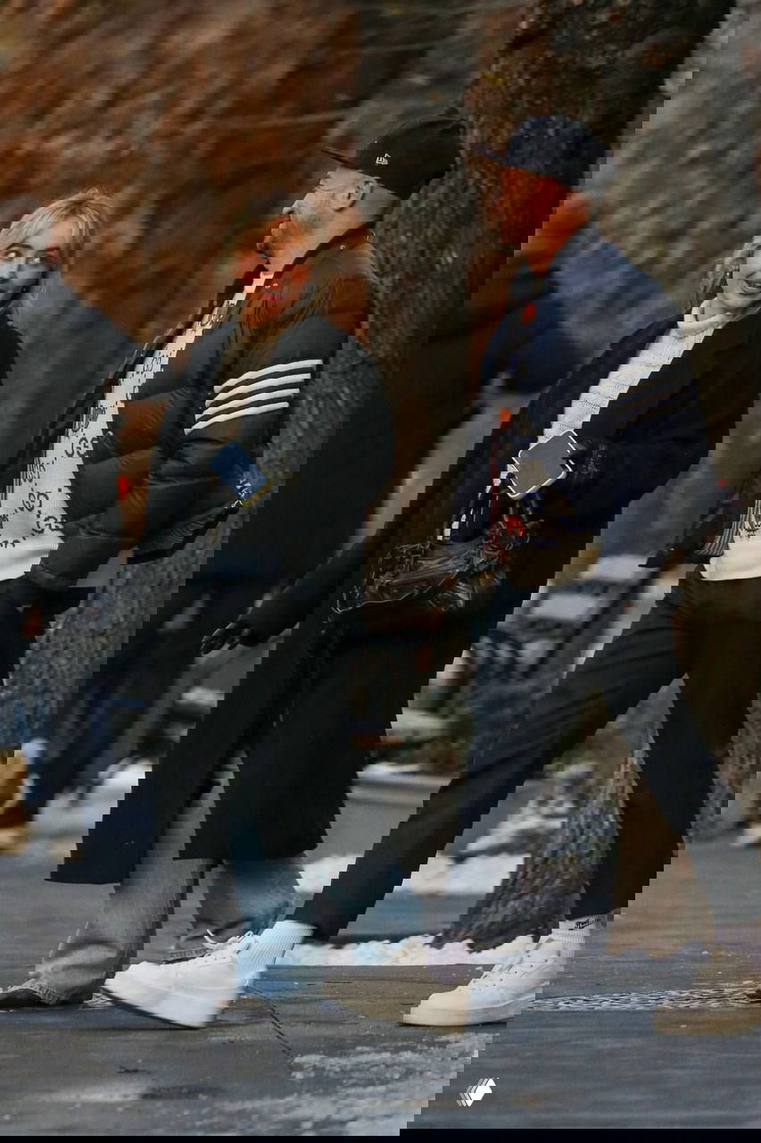 Пара идёт по заснеенному тротуару; женщина в тёмном пальто, джинсах и белых кроссовках беседует с мужчиной на улице.