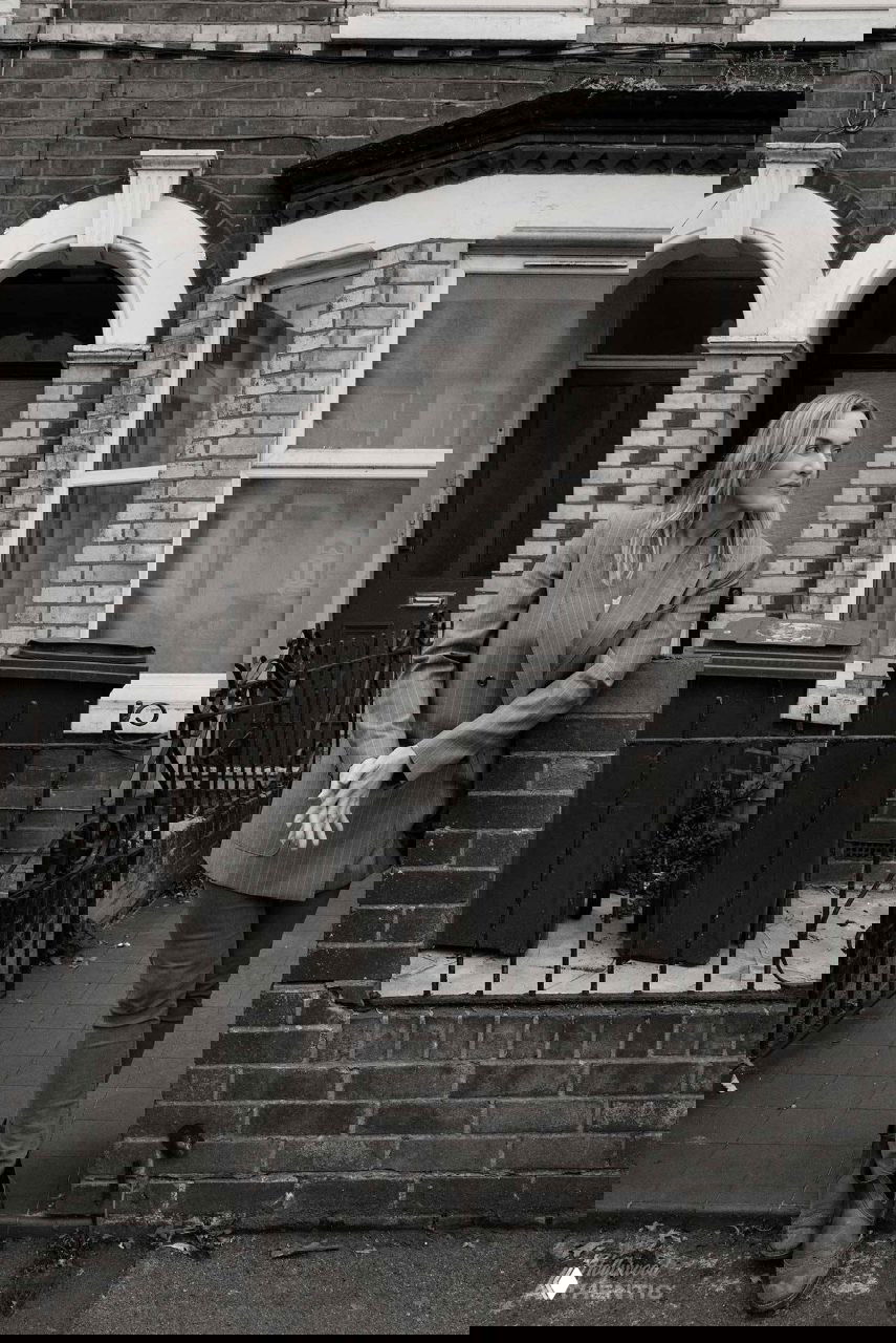 Актриса на тротуаре рядом с типичным жилым домом: чёрно‑белый кадр, передающий атмосферу родного квартала и повседневности.