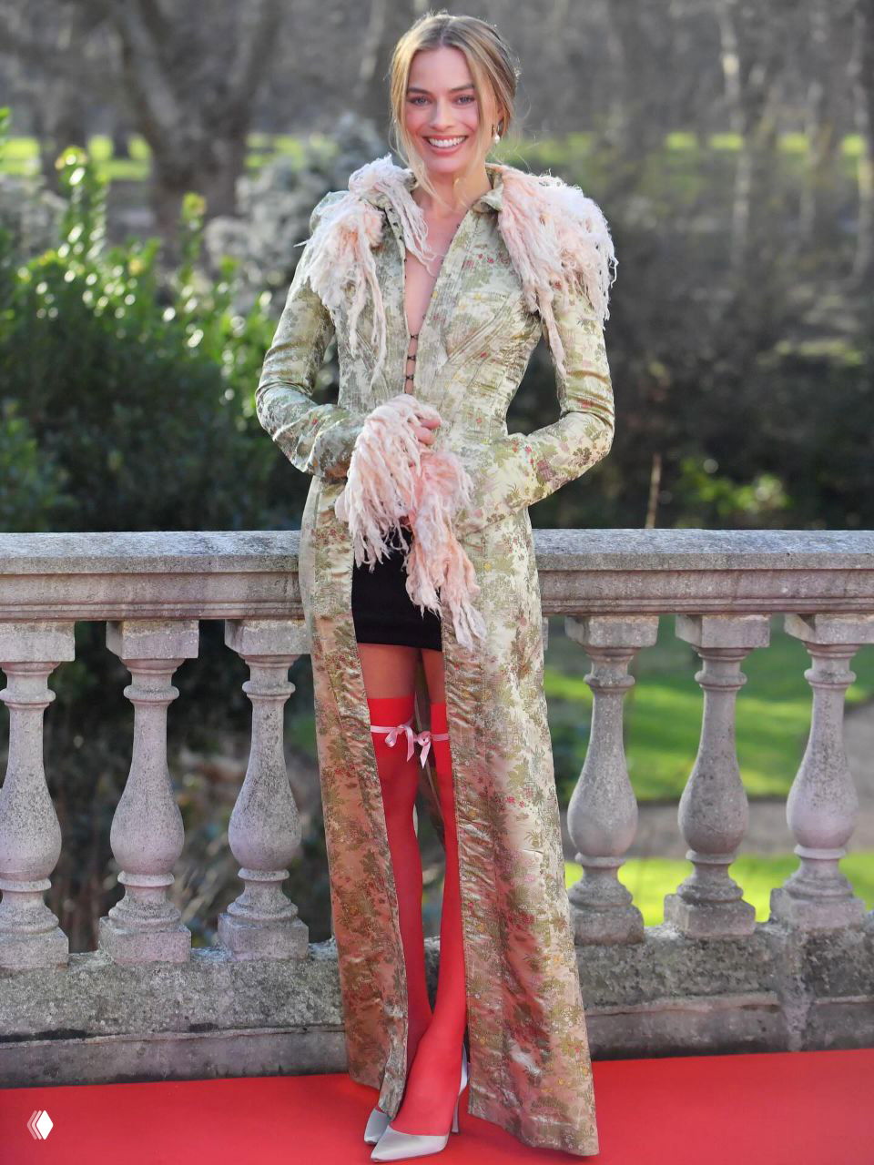 Женщина в длинном фактурном пальто с розовыми перьями и ярко-красными чулками на красной дорожке, полный рост и фон парка