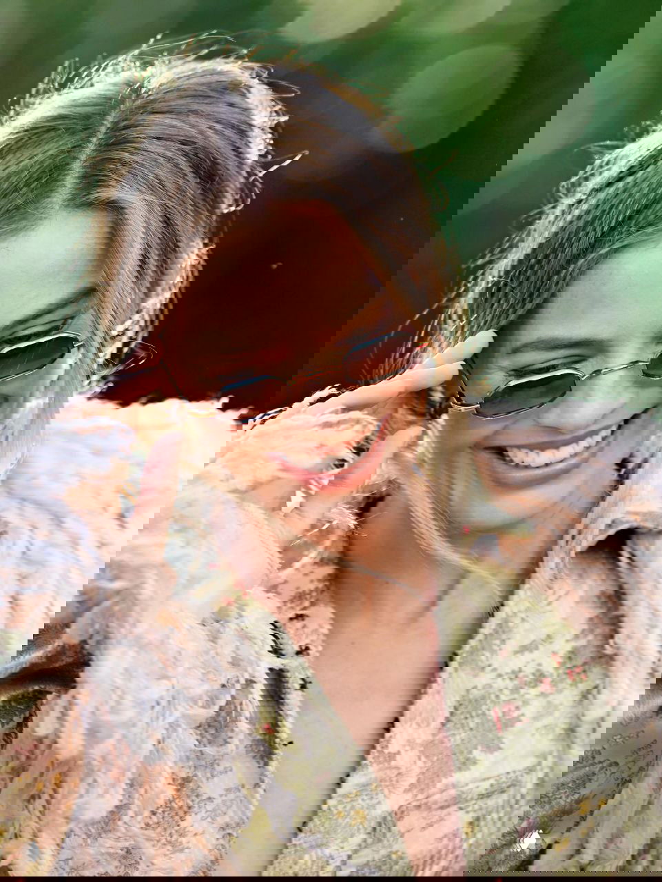 Крупный план улыбающейся женщины в миниатюрных солнечных очках, мягкое боке и естественный макияж на фоне зелёной листвы