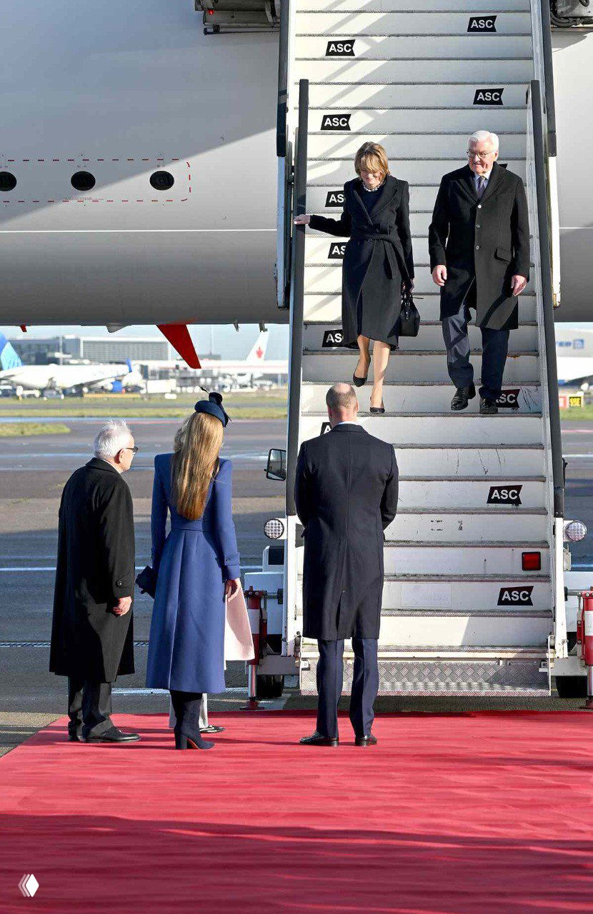 Гости спускаются по трапу самолёта и выходят на красную дорожку в аэропорту Хитроу; женщина в синем пальто стоит на переднем плане.