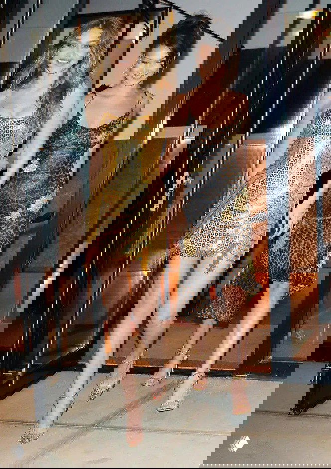 Две женщины в вечерних платьях — одно золотое мини с пайетками и второе в леопардовом шелке — выходят из здания в обуви на высоком каблуке, уличная фэшн-сцена.