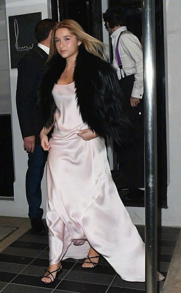 Молодая девушка в светлом атласном платье и тёмной накидке идёт по порогу, её образ привлекает внимание фотографов на афтерпати.