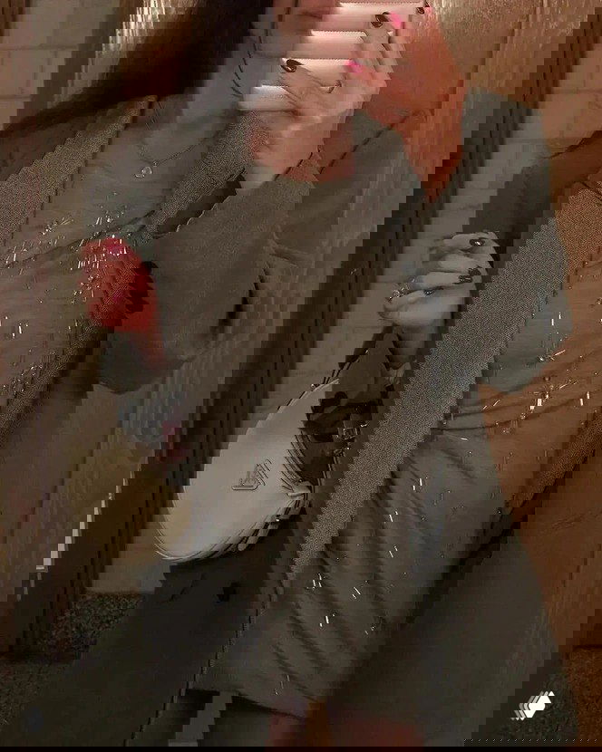 Селфи в зеркале: женщина в сером пальто и платье с металлическими декоративными элементами держит светлую сумку и красит ногти в глубокий красный, интерьер ванной/прихожей.