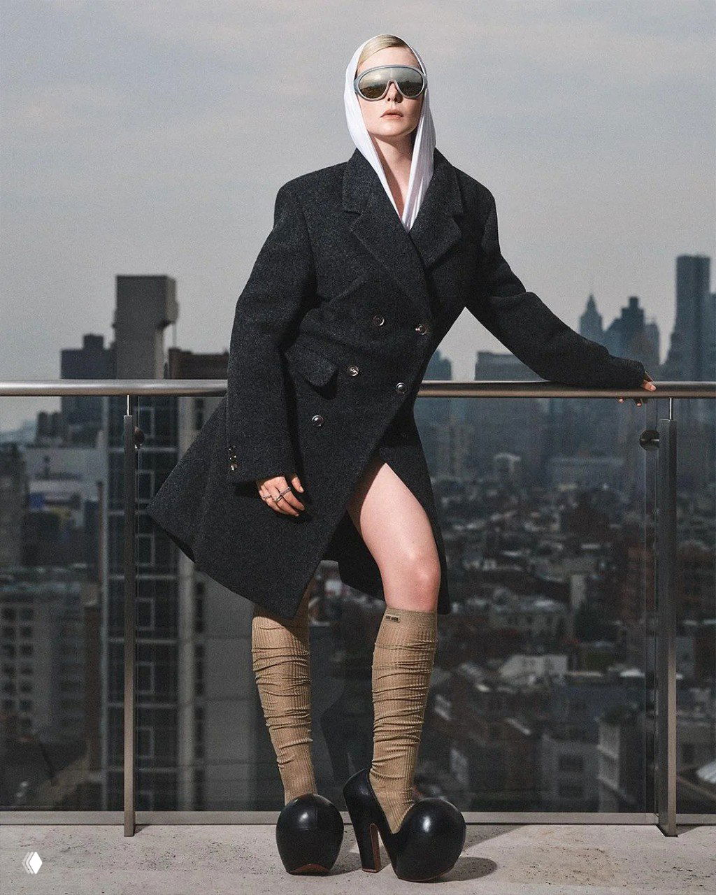 Эль Фаннинг в тёплом двубортном пальто и капюшоне, опирается о перила террасы: строгость кроя и силуэт в городском антураже.