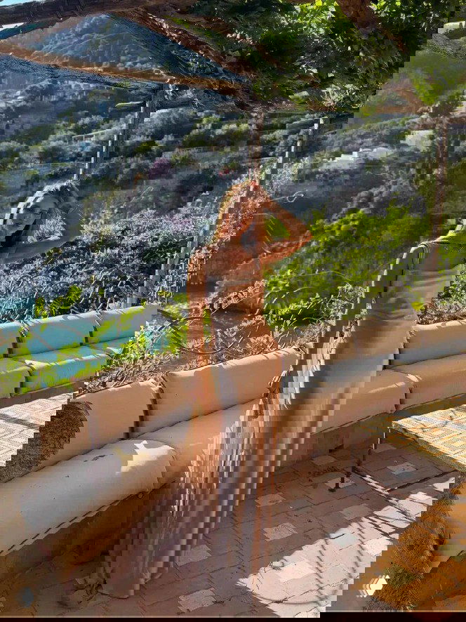 Женщина в длинном струящемся атласном платье позирует на террасе с видом на средиземноморский пейзаж, зелёные склоны и море на заднем плане, солнечная съёмка на открытом воздухе.