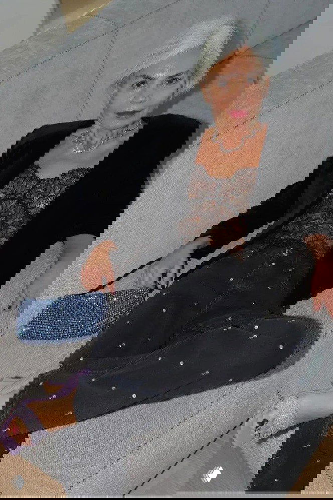 Пожилая женщина с седыми волосами в кружевном топе, коротком пиджаке и черных джинсах с блёстками отдыхает на диване рядом с маленькой сияющей сумочкой, кадр стилизован под модную съёмку.