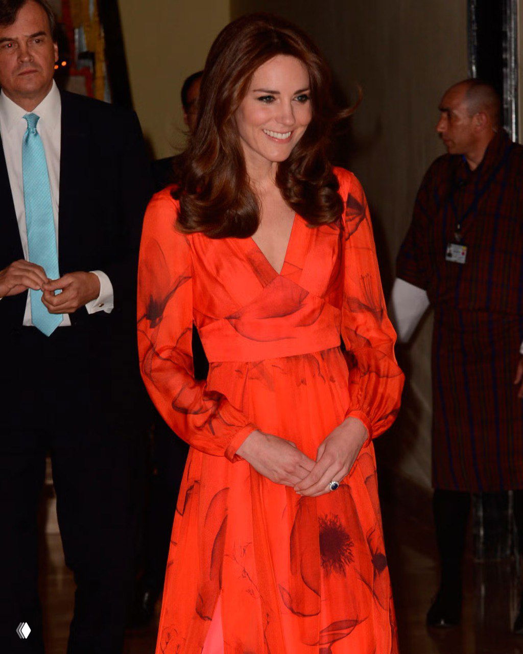 Женщина в ярком красно-оранжевом платье с принтом в интерьере, улыбка и спокойная постановка, фон с людьми в тени.