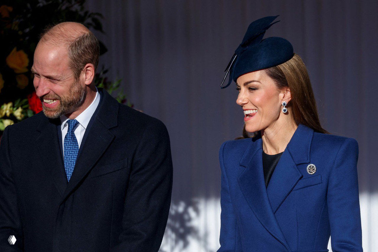 Крупный портретный кадр пары гостей на приёме: женщина в синем пальто с шляпой улыбается рядом с мужчиной в строгом костюме.