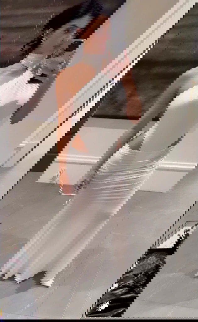 Женщина в белом длинном платье с вырезами делает селфи у стены; образ подчёркивает силуэт и вечерний дресс-код.