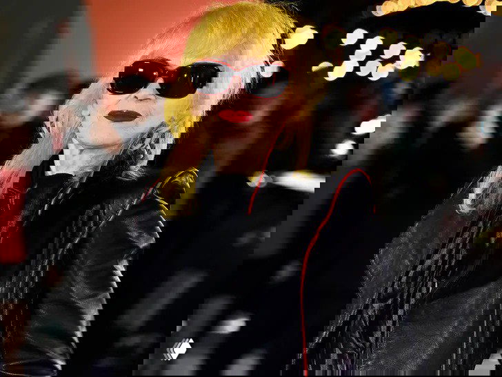 Портрет Пэм Хогг на публичном мероприятии: дизайнер в тёмной кожаной куртке и солнцезащитных очках, с яркой помадой и рок-образом.