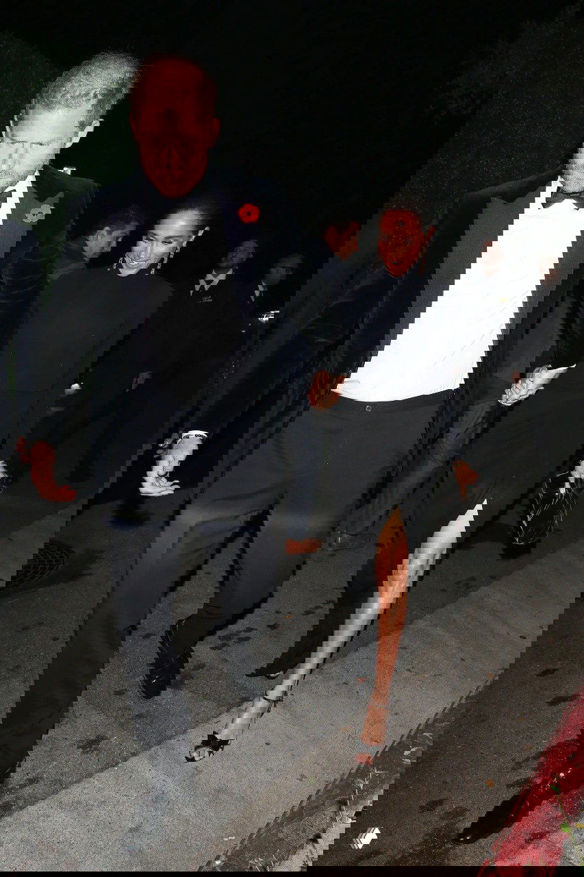 Пара в вечерних нарядах идёт по тротуару ночью: мужчина в смокинге, женщина в чёрном платье с разрезом, держатся за руки.