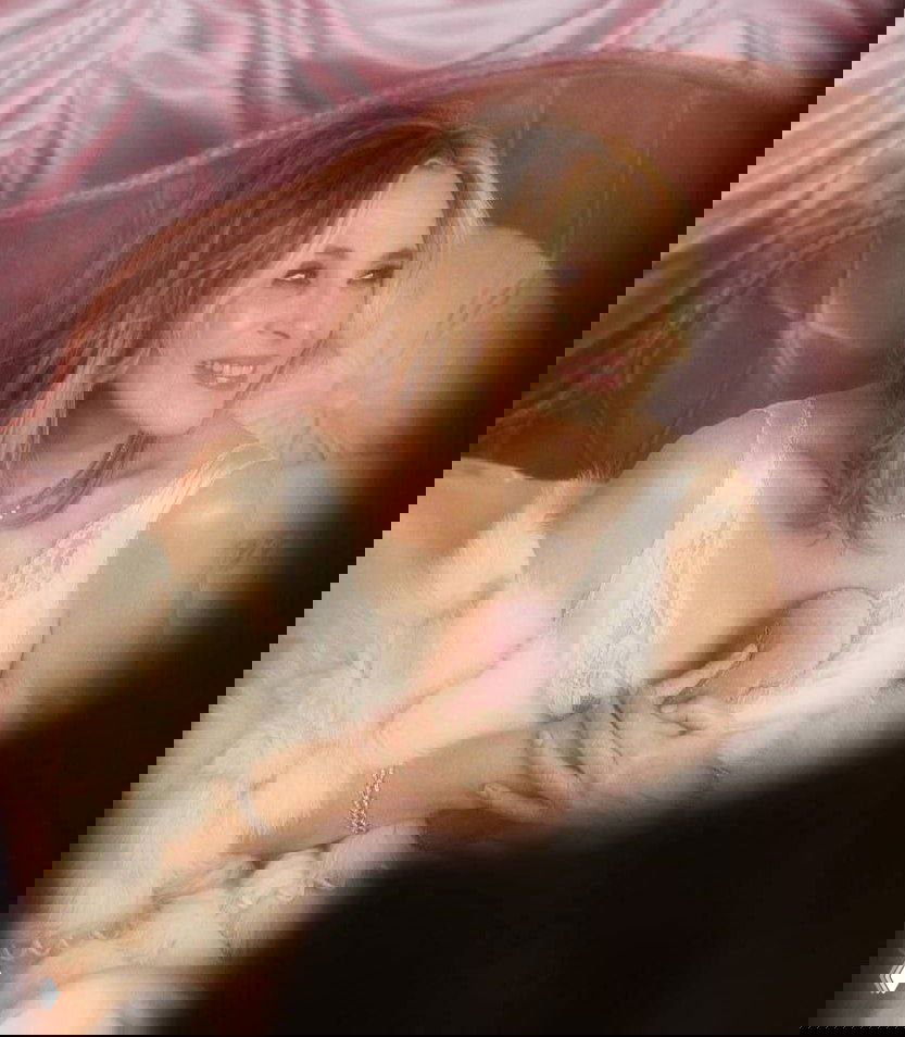 Портретная студийная фотография: женщина в светлом кружевном платье и пушистом шарфе на розовом фоне, мягкий теплый свет.