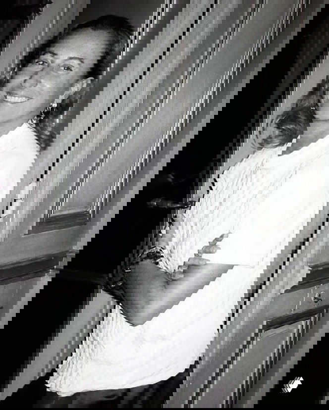 Чёрно-белый портрет улыбающейся женщины в домашних интерьерах — естественный кадр с расслабленной позой у двери, атмосферный снимок.