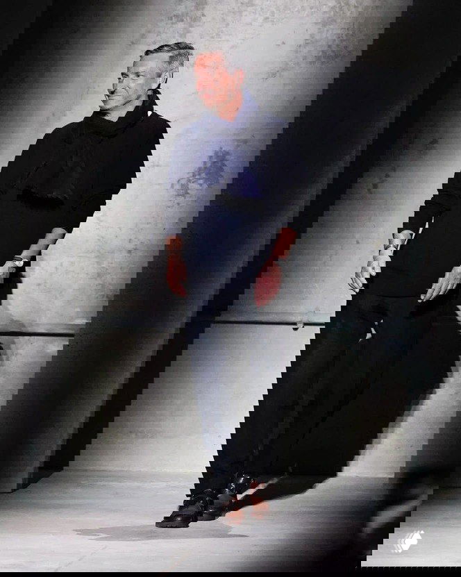 Фронтальный снимок мужчины в темной одежде на подиуме или в студии — полный рост, уверенная поза и деловой образ в нейтральном фоне.