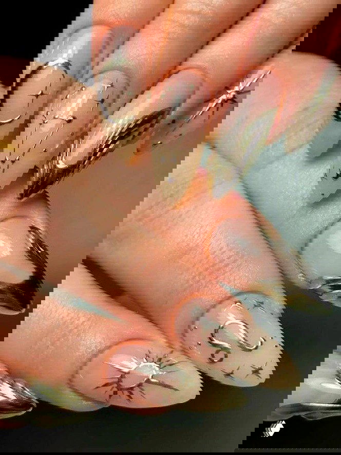 Острые миндалевидные ногти нюдового оттенка с золотыми металлическими символами — месяц, созвездие и тонкие акценты, аккуратный глянцевый финиш.