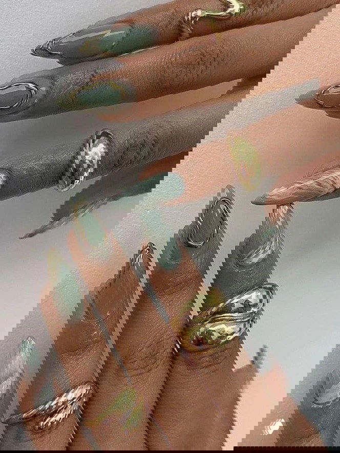 Мраморный зелёно‑мятный маникюр с золотыми объёмными рамками и глянцевым финишем на длинных ногтях, дополненный крупными золотыми кольцами на пальцах.