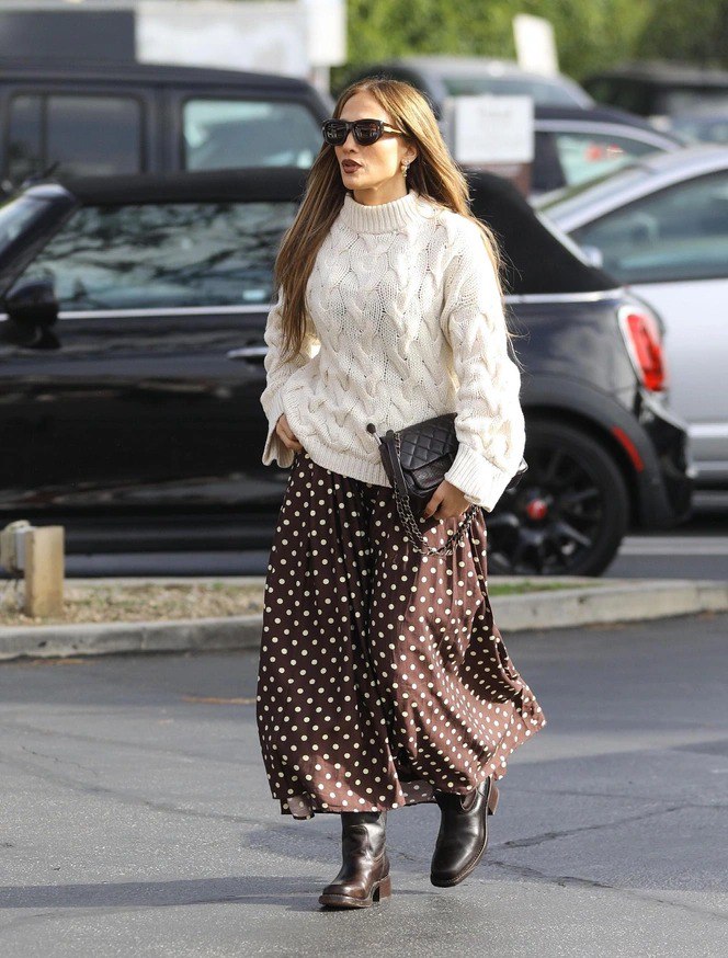 Крупный план: образ Дженнифер Лопес — объёмный белый свитер, длинное коричневое платье в горох, тёмная сумка через руку и солнцезащитные очки.