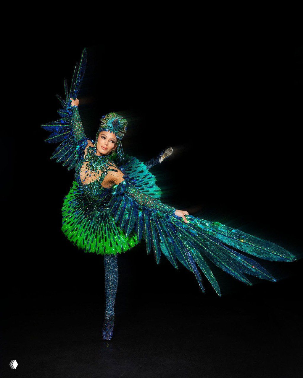 Участница в ярком зелёно‑синем национальном костюме с большими крыльями и перьевыми деталями на тёмном фоне, позирует.