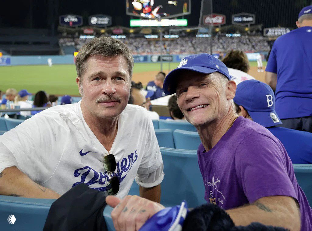 Два зрителя на трибунах бейсбольного матча: мужчина в белой рубашке и сосед в синей кепке, на заднем плане поле и трибуны стадиона.