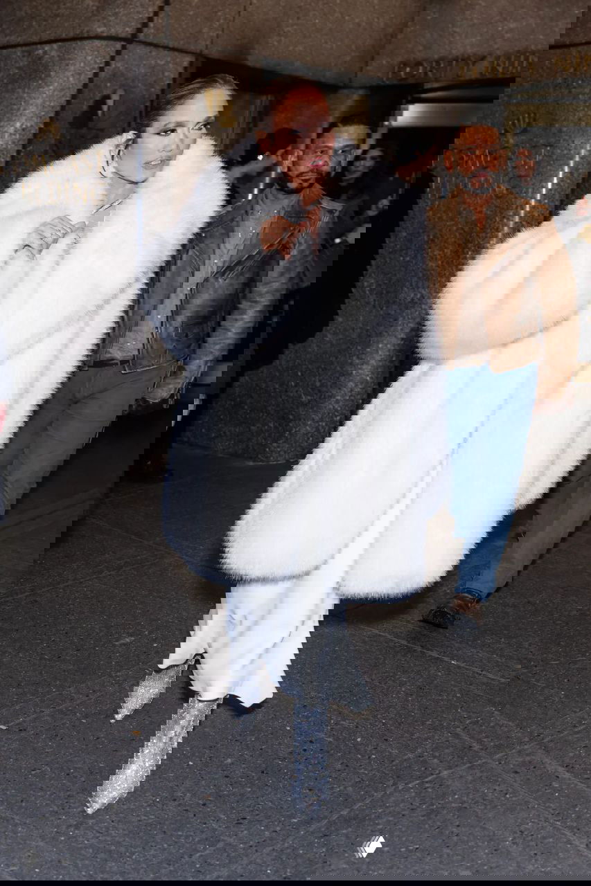 Женщина в длинной белой пушистой шубе и блестящих ботильонах идёт по тротуару вечером, вокруг прохожие и фотографы.