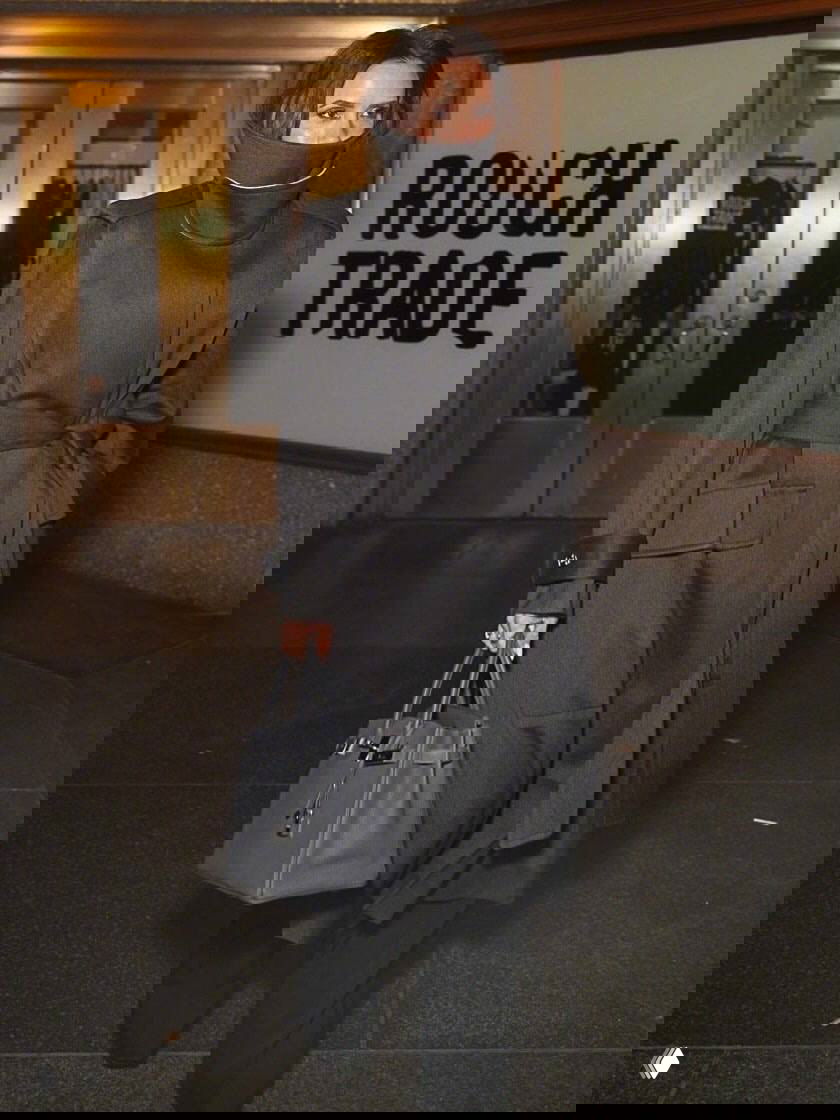 Женщина в тёмном объёмном пальто и широким воротником выходит из здания с сумкой в руке на вечерней улице, спокойный классический образ