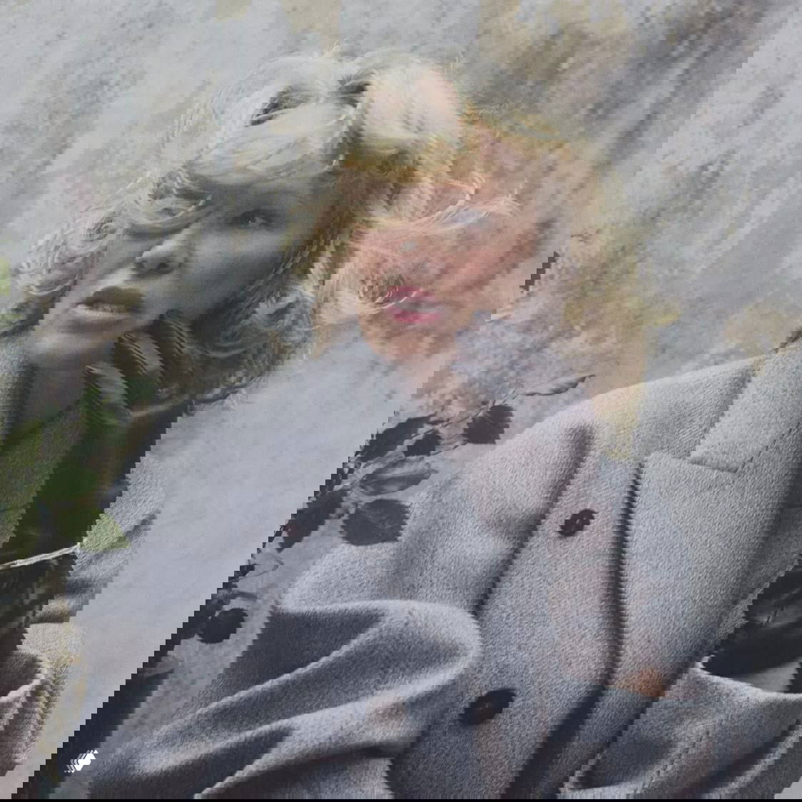 Портрет Памелы Андерсон в сером пальто и чёрных перчатках на фоне потертой стены — кадр из глянцевой фотосессии.