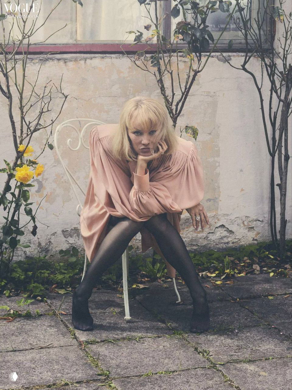 Памела Андерсон сидит на металлическом кресле в нежно-розовом платье и чёрных колготках, задумчивый образ для журнала.
