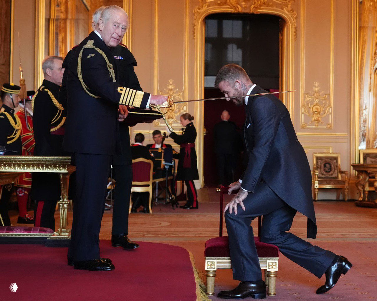 Король Карл посвящает Дэвида Бекхэма в рыцари в зале Виндзорского замка: Бекхэм на колене, меч над его плечом в торжественной обстановке.