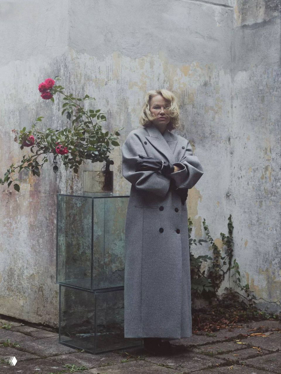 Полнофигурный кадр Памелы Андерсон в длинном сером пальто рядом с вазой и цветами на старом бетонном фоне.