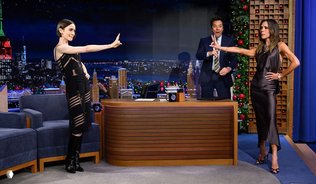 Сцена ночного ток‑шоу с ведущим и двумя гостьями; одна демонстрирует жест, другая стоит рядом в шёлковом платье — студийная сценография в кадре