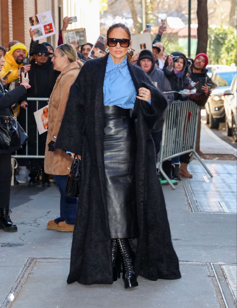 Длинная чёрная шуба поверх голубой рубашки и кожаной юбки, городской фон с собравшимися фанатами и фотографами.