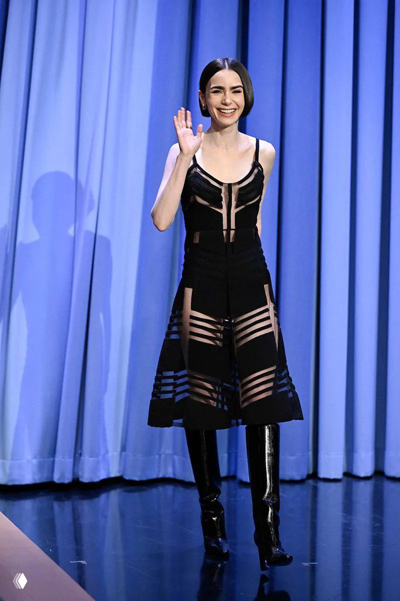 Женщина на сцене в чёрном платье с геометричными прозрачными элементами и высокими лаковыми ботфортами, приветствует публику на шоу