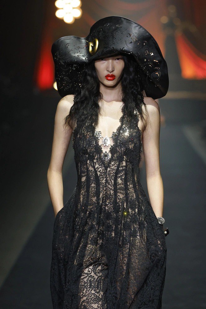 Подиумный кадр: модель в чёрном кружевном платье и крупной шляпе, яркая красная помада подчёркивает губы и создаёт драматичный вечерний образ на шоу