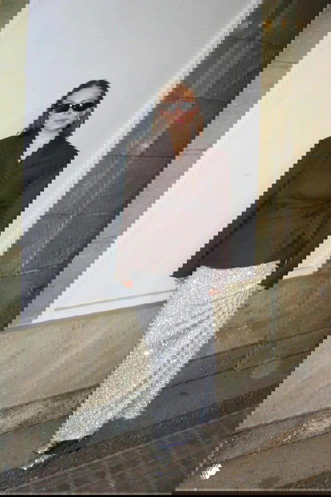 Женский праздничный образ: тёмный приталенный жакет в сочетании с длинной серебристой блестящей юбкой, солнцезащитные очки, уличная стена на заднем плане.