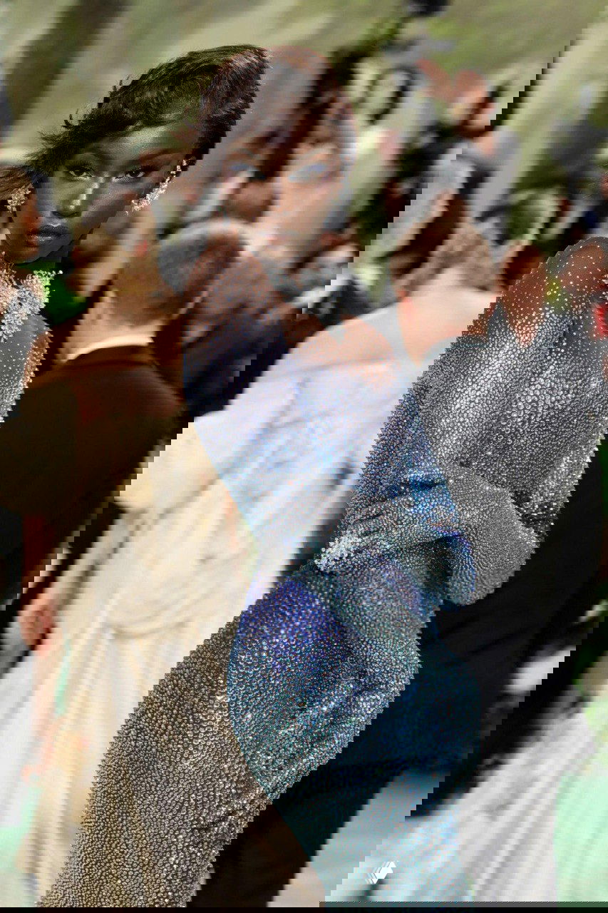 Темнокожая модель в сверкающем голубом платье и украшениях позирует на красной дорожке на фоне фотографов и гостей.