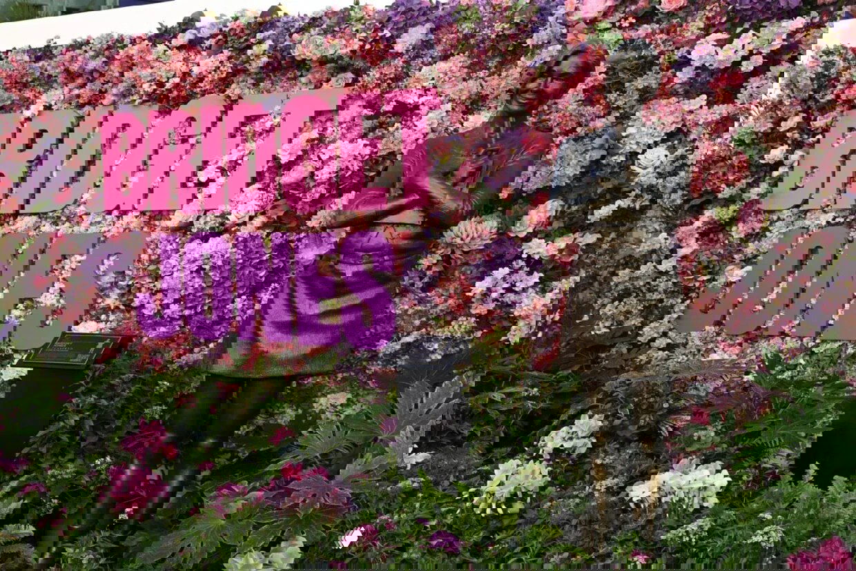 Бронзовая статуя Бриджит Джонс на фоне плотной цветочной стены с большими розово‑фиолетовыми буквами BRIDGET JONES в Лондоне.