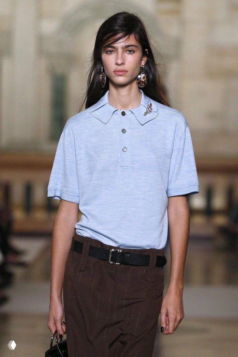 Модель на подиуме в светлом топе с длинными волосами и косой чёлкой; образ в духе подиумных показов, минимальный макияж.