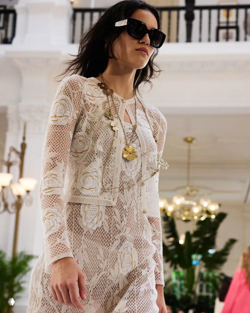 Крупный план модели в кружевном наряде Chanel с длинными цепочками и крупными солнцезащитными очками на фоне люстр и растений на показе.