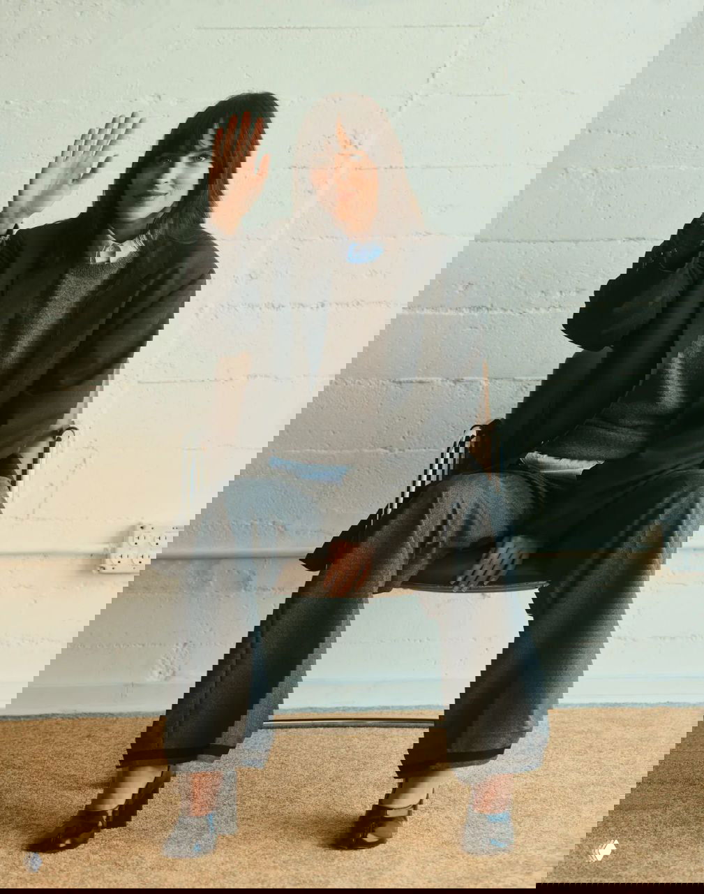 Портретная кадрировка актрисы в просторном костюме и туфлях на небольшом стуле; интерьерная съёмка с нейтральной стеной и ретро-атмосферой.